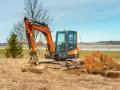 Pomarańczowa minikoparka na gąsienicach stoi na zaoranym polu, obok usypana kopa ziemi. Czas na wymianę oleju hydraulicznego w minikoparce.