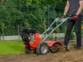 Osoba w rękawiczkach obsługuje czerwoną glebogryzarkę spalinową, spulchniając ziemię w ogrodzie. Dowiedz się, jak używać glebogryzarki spalinowej.