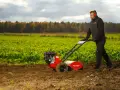 Mężczyzna w słuchawkach ochronnych pracuje glebogryzarką na polu. Jesienne drzewa w tle.
