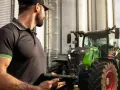 Rolnik z tabletem przy silosach, obok zielony traktor Fendt. W tle biała furgonetka.
