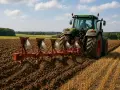 Zielony traktor z pługiem wykonuje oranie ziemi na polu. W tle widać las i błękitne niebo z chmurami.