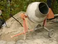 Pomarańczowa betoniarka 250l gotowa do pracy, obok sterta piasku i łopaty.
