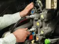 Ręce mechanika pracują przy skomplikowanym układzie hydraulicznym. Widać węże i zawory z kolorowymi oznaczeniami.