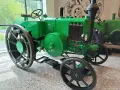 Zielony, zabytkowy Ursus, najstarszy model, z wielkimi kołami i kierownicą, stoi w muzeum.