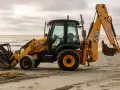 Żółta koparko-ładowarka firmy JCB stoi na piaszczystej plaży, z widokiem na morze i zachmurzone niebo.