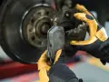 Mechanik w żółtych rękawicach wymienia klocki hamulcowe. Widoczny jest tarczowy układ hamulcowy.