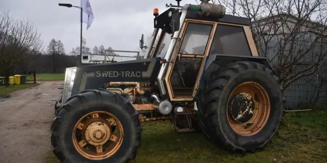 Ciemny traktor SWED-TRAC z pomarańczowymi światłami ostrzegawczymi i dużymi kołami stoi na trawie.