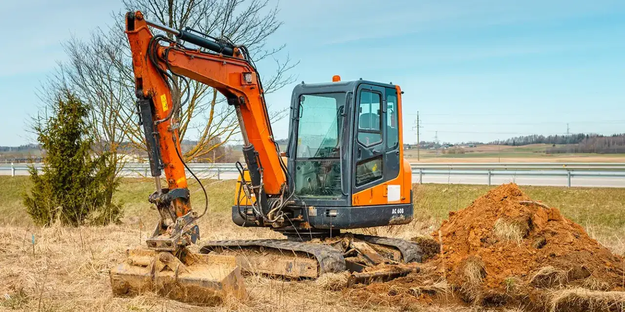 Pomarańczowa minikoparka na gąsienicach stoi na zaoranym polu, obok usypana kopa ziemi. Czas na wymianę oleju hydraulicznego w minikoparce.