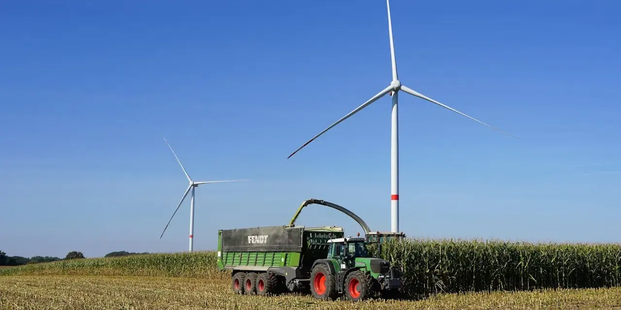 Zielone Fendt 310 z przyczepą zbiera kukurydzę pod wiatrakami. Dane techniczne tej maszyny pozwalają na wydajną pracę.