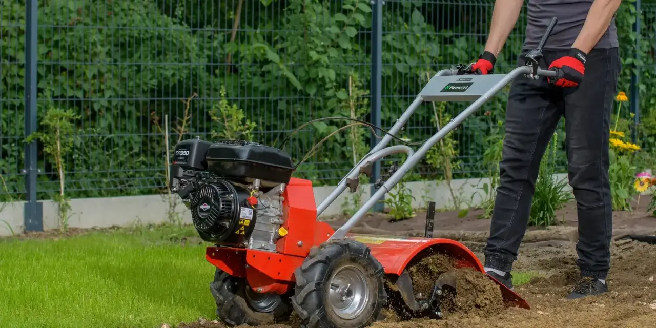 Osoba w rękawiczkach obsługuje czerwoną glebogryzarkę spalinową, spulchniając ziemię w ogrodzie. Dowiedz się, jak używać glebogryzarki spalinowej.