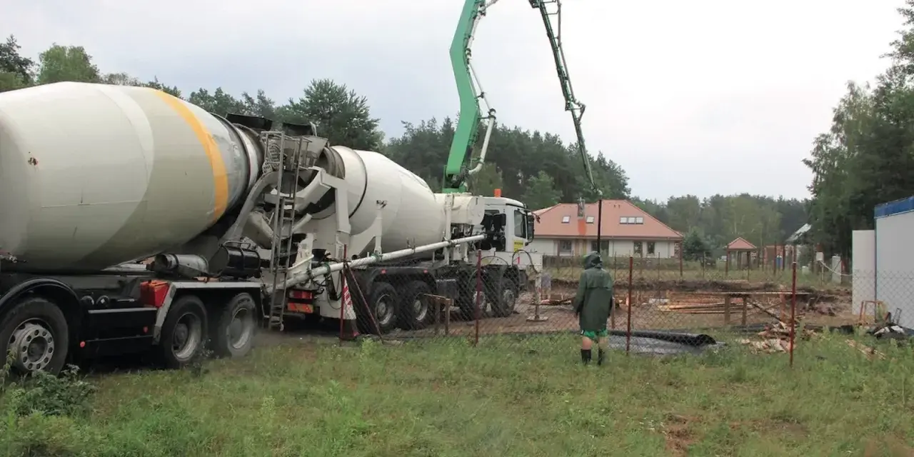 Betoniarka z wysięgnikiem pompuje beton. Pracownik w zielonej kurtce obserwuje. To ważny etap budowy, a czyszczenie betoniarki po pracy, jak usunąć zaschnięty beton z betoniarki, jest kluczowe.