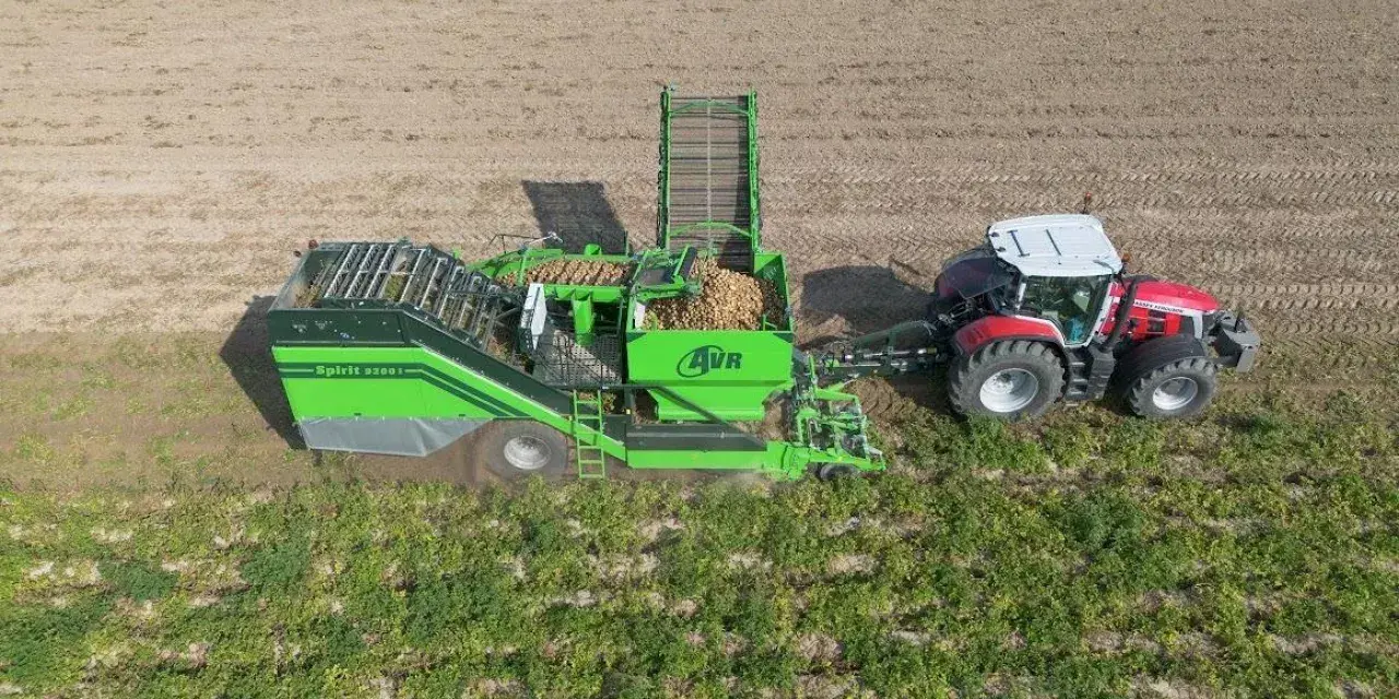 Kombajn AVR Spirit 9200 zbiera ziemniaki na polu. Czerwony traktor ciągnie maszynę, która wypełnia się plonami.