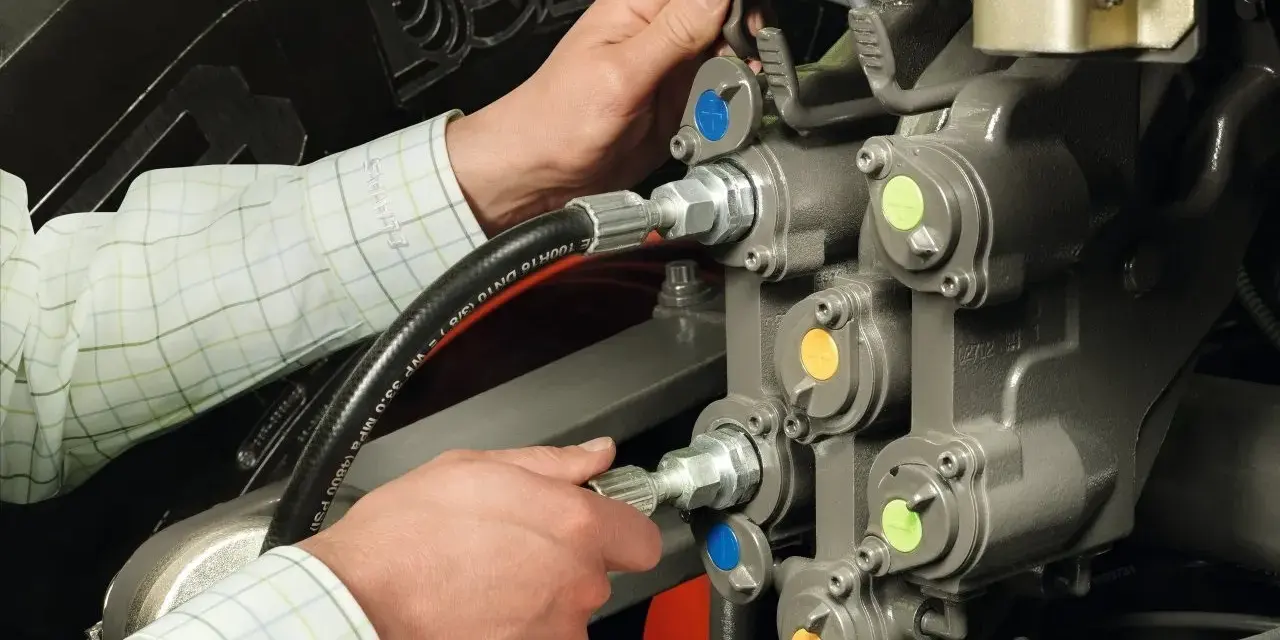 Ręce mechanika pracują przy skomplikowanym układzie hydraulicznym. Widać węże i zawory z kolorowymi oznaczeniami.