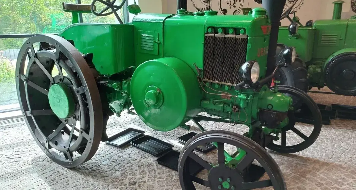 Zielony, zabytkowy Ursus, najstarszy model, z wielkimi kołami i kierownicą, stoi w muzeum.