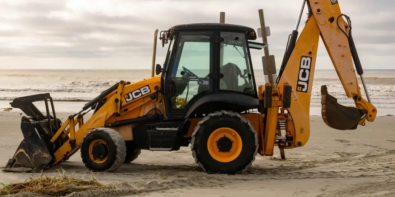 Żółta koparko-ładowarka firmy JCB stoi na piaszczystej plaży, z widokiem na morze i zachmurzone niebo.