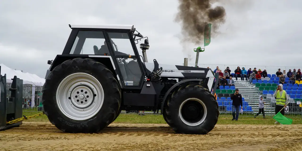 Ciężki traktor Ursus 1224 z potężnym dymem z rury wydechowej podczas zawodów tractor pulling. Widownia obserwuje z trybun.