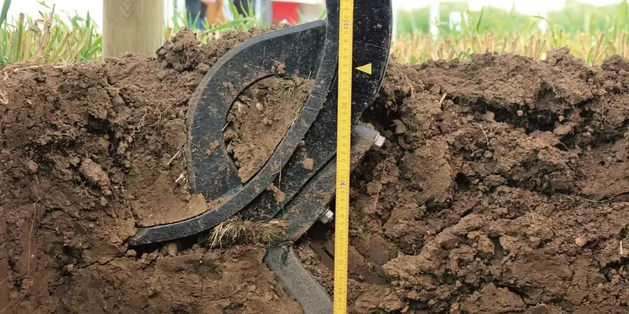 Czarne, metalowe elementy podeszwy płużnej zagłębiają się w ziemi. Obok miarka pokazuje głębokość orki.