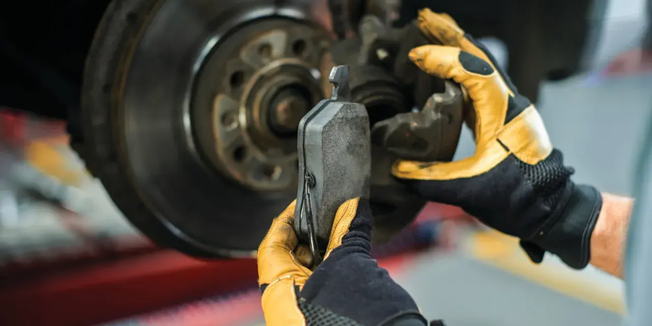 Mechanik w żółtych rękawicach wymienia klocki hamulcowe. Widoczny jest tarczowy układ hamulcowy.