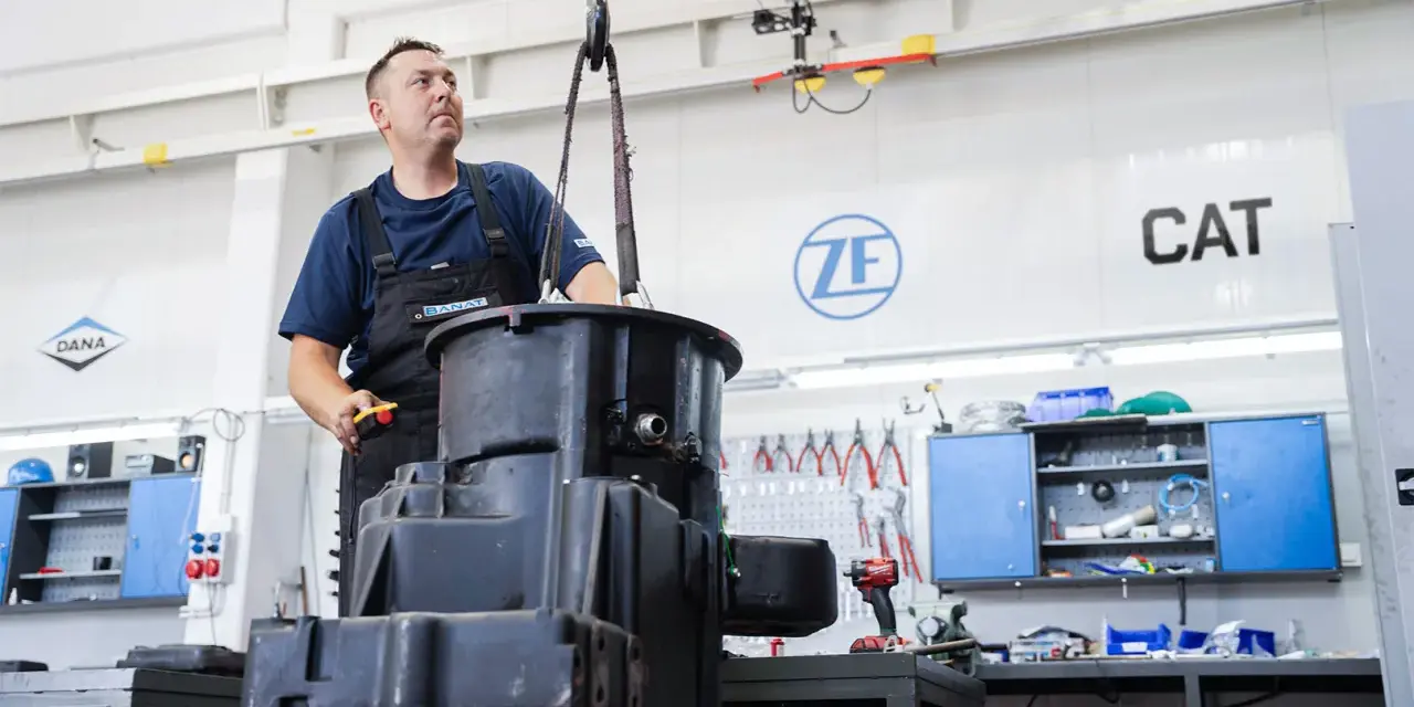 Mechanik steruje podnoszeniem dużych elementów skrzyni biegów w warsztacie. W tle logo ZF i CAT.