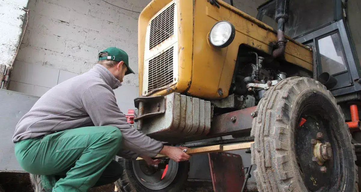 Mężczyzna w zielonych spodniach i czapce reguluje przednie koło żółtego ciągnika 4x4, pokazując, jak ustawić zbieżność.