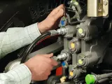 Ręce mechanika pracują przy skomplikowanym układzie hydraulicznym. Widać węże i zawory z kolorowymi oznaczeniami.