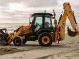 Żółta koparko-ładowarka firmy JCB stoi na piaszczystej plaży, z widokiem na morze i zachmurzone niebo.