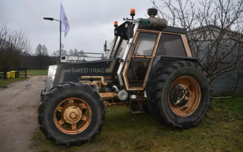 Ciemny traktor SWED-TRAC z pomarańczowymi światłami ostrzegawczymi i dużymi kołami stoi na trawie.
