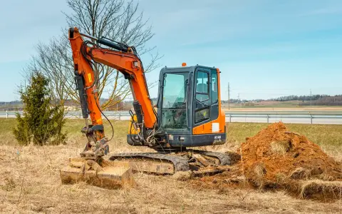 Pomarańczowa minikoparka na gąsienicach stoi na zaoranym polu, obok usypana kopa ziemi. Czas na wymianę oleju hydraulicznego w minikoparce.