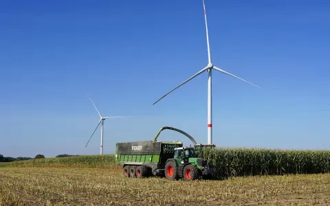 Zielone Fendt 310 z przyczepą zbiera kukurydzę pod wiatrakami. Dane techniczne tej maszyny pozwalają na wydajną pracę.
