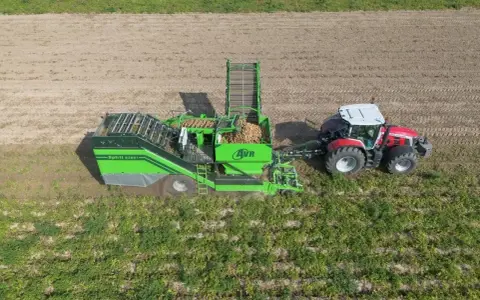 Kombajn AVR Spirit 9200 zbiera ziemniaki na polu. Czerwony traktor ciągnie maszynę, która wypełnia się plonami.