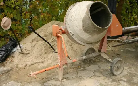 Pomarańczowa betoniarka 250l gotowa do pracy, obok sterta piasku i łopaty.