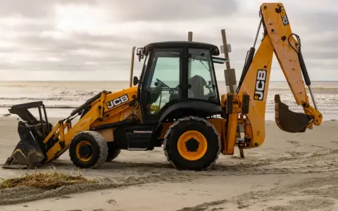 Żółta koparko-ładowarka firmy JCB stoi na piaszczystej plaży, z widokiem na morze i zachmurzone niebo.