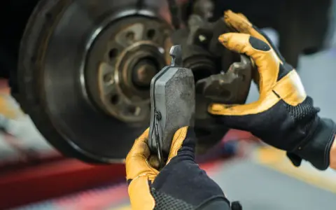 Mechanik w żółtych rękawicach wymienia klocki hamulcowe. Widoczny jest tarczowy układ hamulcowy.
