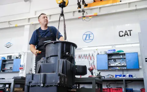 Mechanik steruje podnoszeniem dużych elementów skrzyni biegów w warsztacie. W tle logo ZF i CAT.
