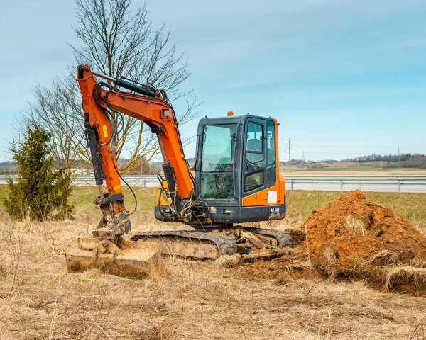 Pomarańczowa minikoparka na gąsienicach stoi na zaoranym polu, obok usypana kopa ziemi. Czas na wymianę oleju hydraulicznego w minikoparce.