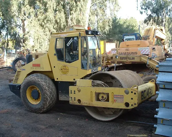 Żółty walec drogowy BOMAG BW 213D-3 gotowy do budowy drogi. W tle koparka Samsung Caterpillar.