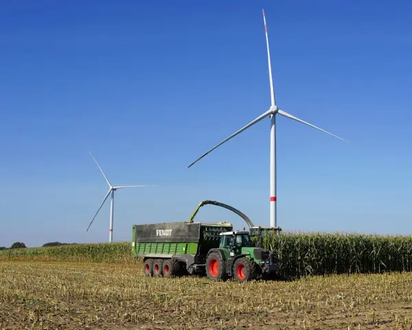 Zielone Fendt 310 z przyczepą zbiera kukurydzę pod wiatrakami. Dane techniczne tej maszyny pozwalają na wydajną pracę.