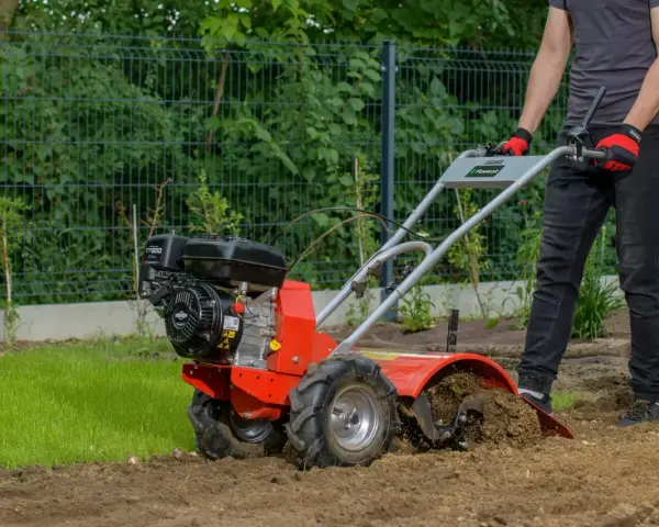 Osoba w rękawiczkach obsługuje czerwoną glebogryzarkę spalinową, spulchniając ziemię w ogrodzie. Dowiedz się, jak używać glebogryzarki spalinowej.