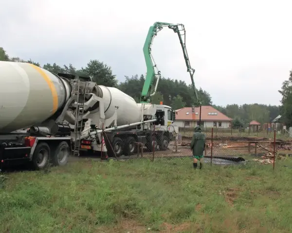 Betoniarka z wysięgnikiem pompuje beton. Pracownik w zielonej kurtce obserwuje. To ważny etap budowy, a czyszczenie betoniarki po pracy, jak usunąć zaschnięty beton z betoniarki, jest kluczowe.