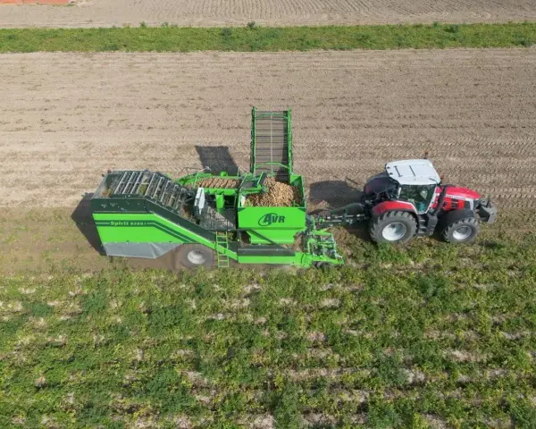 Kombajn AVR Spirit 9200 zbiera ziemniaki na polu. Czerwony traktor ciągnie maszynę, która wypełnia się plonami.
