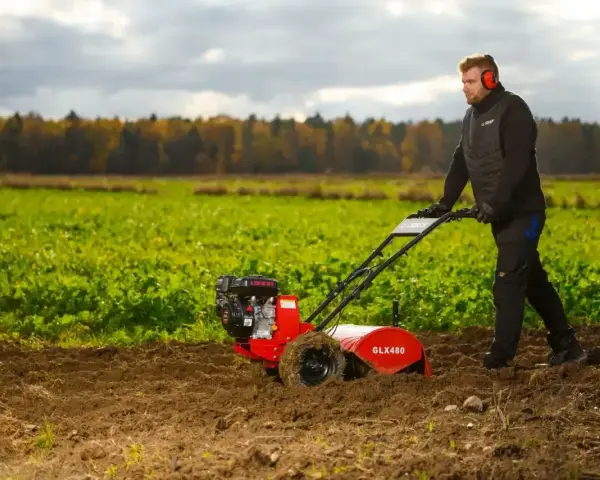 Mężczyzna w słuchawkach ochronnych pracuje glebogryzarką na polu. Jesienne drzewa w tle.