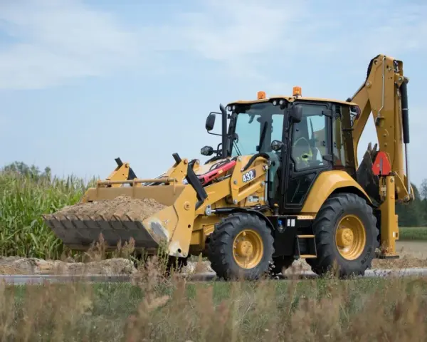 Żółta koparko-ładowarka CAT 432 w trakcie budowy drogi, z pełną łyżką ziemi.