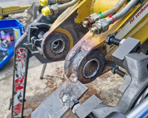 Naprawa tulejowania koparki. Widoczne elementy hydrauliczne i zardzewiałe sworznie.