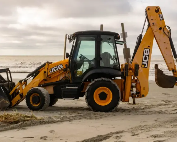 Żółta koparko-ładowarka firmy JCB stoi na piaszczystej plaży, z widokiem na morze i zachmurzone niebo.