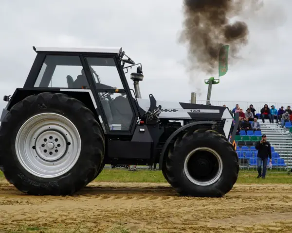 Ciężki traktor Ursus 1224 z potężnym dymem z rury wydechowej podczas zawodów tractor pulling. Widownia obserwuje z trybun.