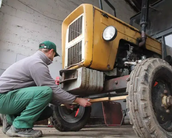 Mężczyzna w zielonych spodniach i czapce reguluje przednie koło żółtego ciągnika 4x4, pokazując, jak ustawić zbieżność.