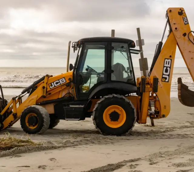 Żółta koparko-ładowarka firmy JCB stoi na piaszczystej plaży, z widokiem na morze i zachmurzone niebo.
