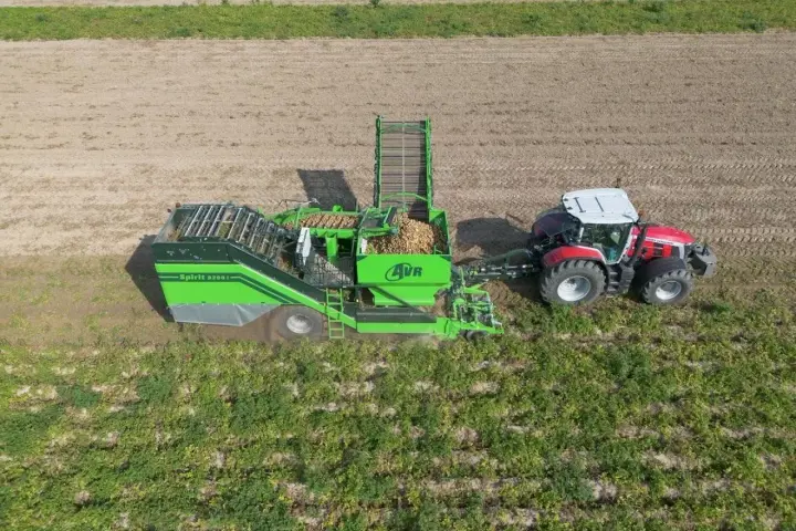 Kombajn AVR Spirit 9200 zbiera ziemniaki na polu. Czerwony traktor ciągnie maszynę, która wypełnia się plonami.