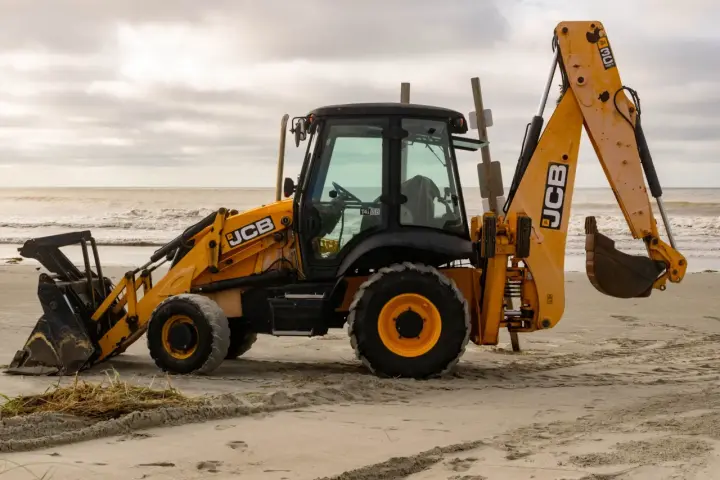 Żółta koparko-ładowarka firmy JCB stoi na piaszczystej plaży, z widokiem na morze i zachmurzone niebo.