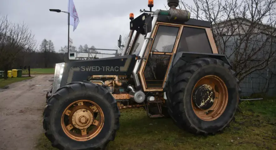 Ciemny traktor SWED-TRAC z pomarańczowymi światłami ostrzegawczymi i dużymi kołami stoi na trawie.